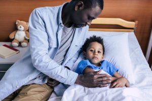 man sitting near sick son lying in bed in hospital
